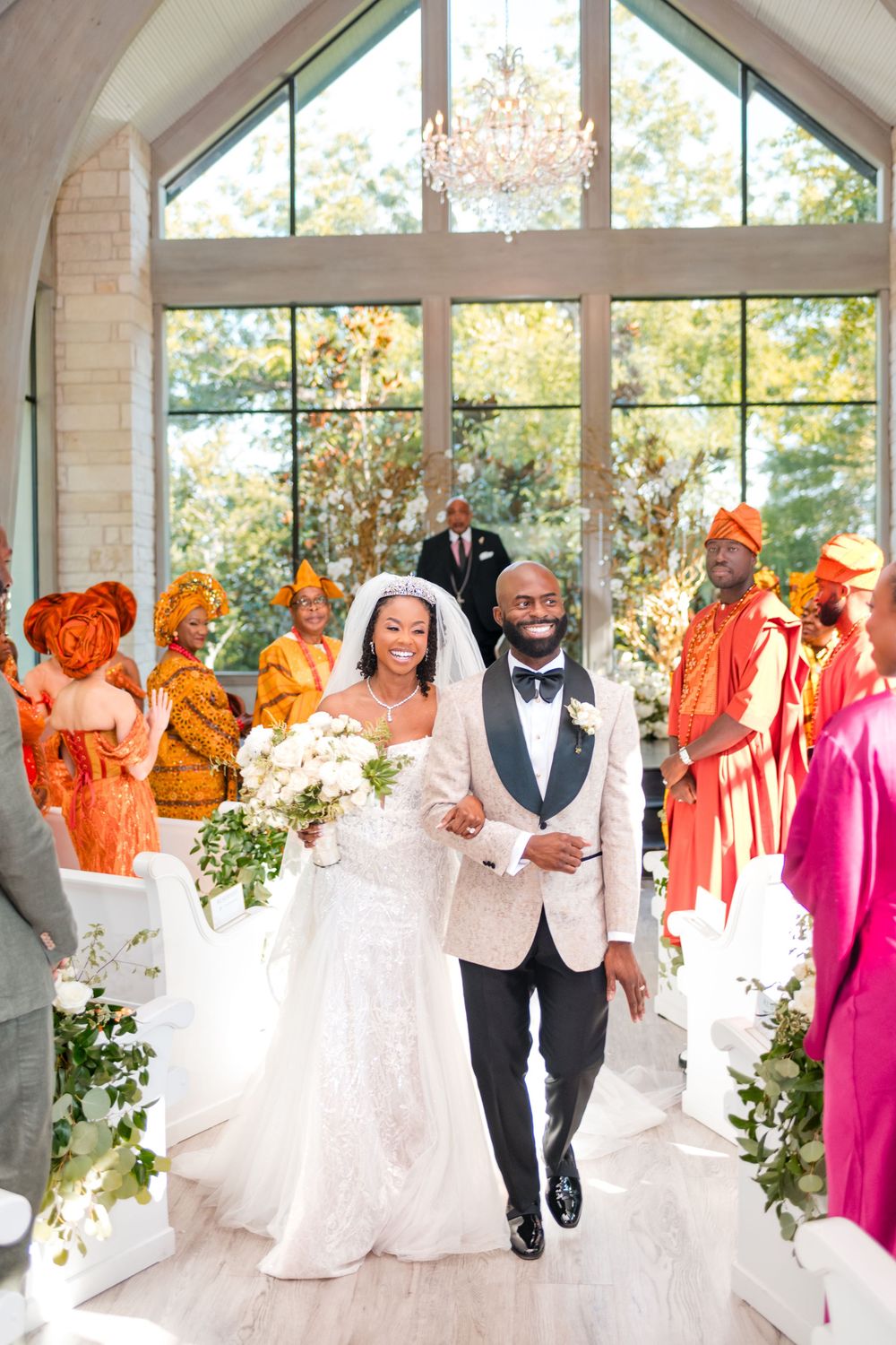 Wedding party walks and dances joyfully in bright formal attire inside a sunlit venue with large windows.