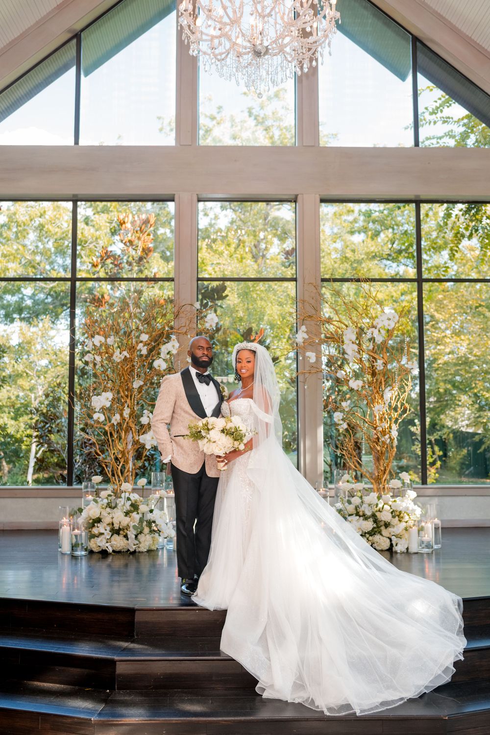 Elegant wedding couple dances in a sunlit venue with floor-to-ceiling windows and floral decorations.