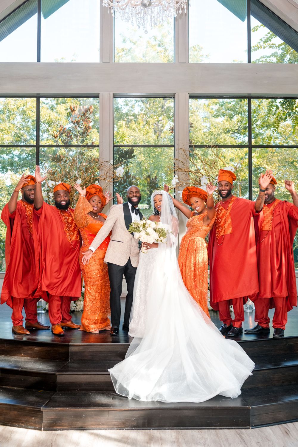 Wedding party in bright orange and red attire poses with bride in white gown against large windows during celebration.