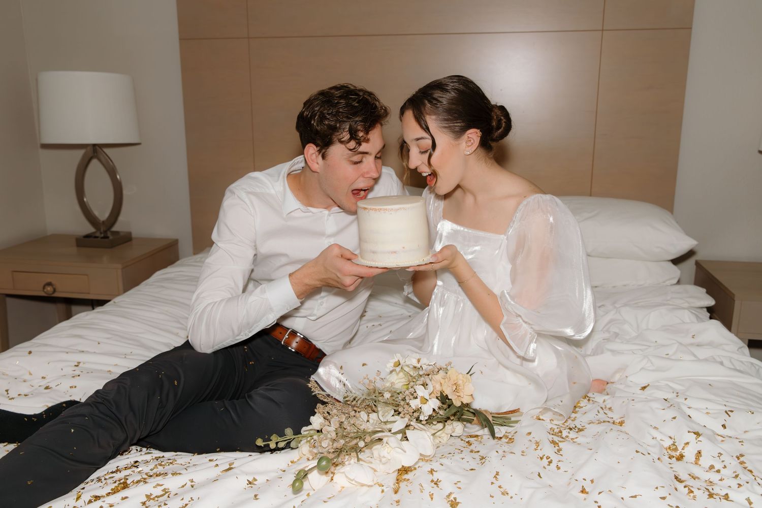 A romantic couple in wedding attire shares intimate moments while lying on a bed covered in flower petals.
