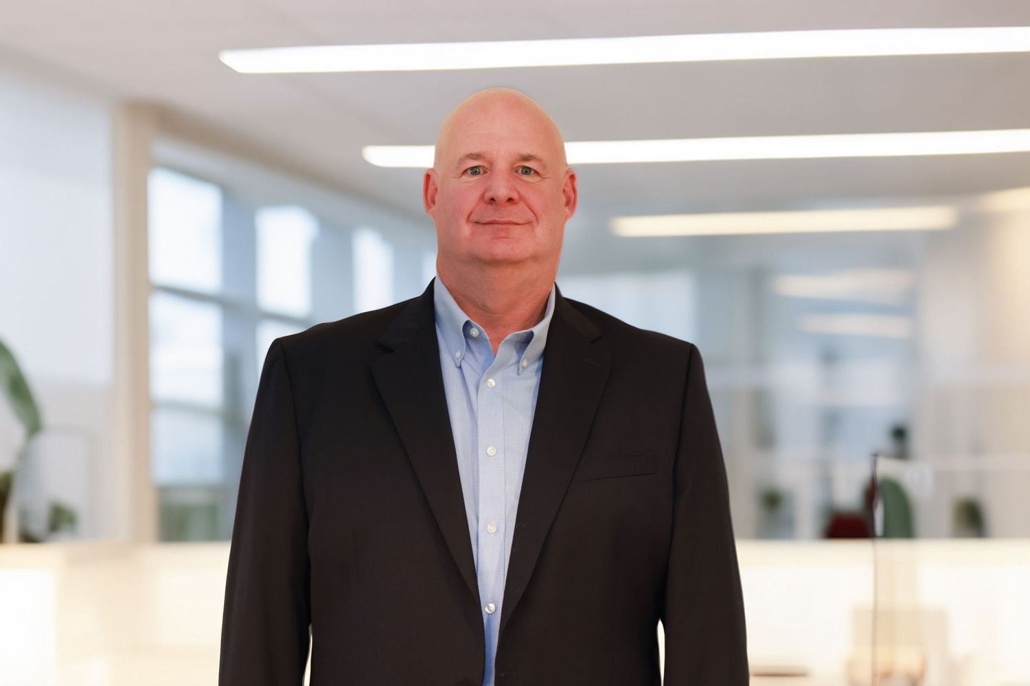 Professional in a dark suit jacket and light blue button-down shirt stands in a bright modern office setting.