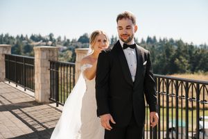 A couple in wedding attire poses on an outdoor balcony with scenic mountain views in the background.