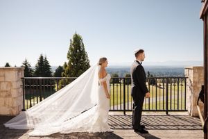 A wedding couple poses together on a scenic balcony with a long flowing veil trailing behind them in the sunlight.