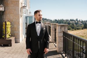 A groom in a classic black tuxedo stands on a stone balcony overlooking scenic hills and trees on a sunny day.