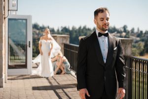 A groom in a black tuxedo stands on a balcony while a bride in white is visible in the background on a sunny day.