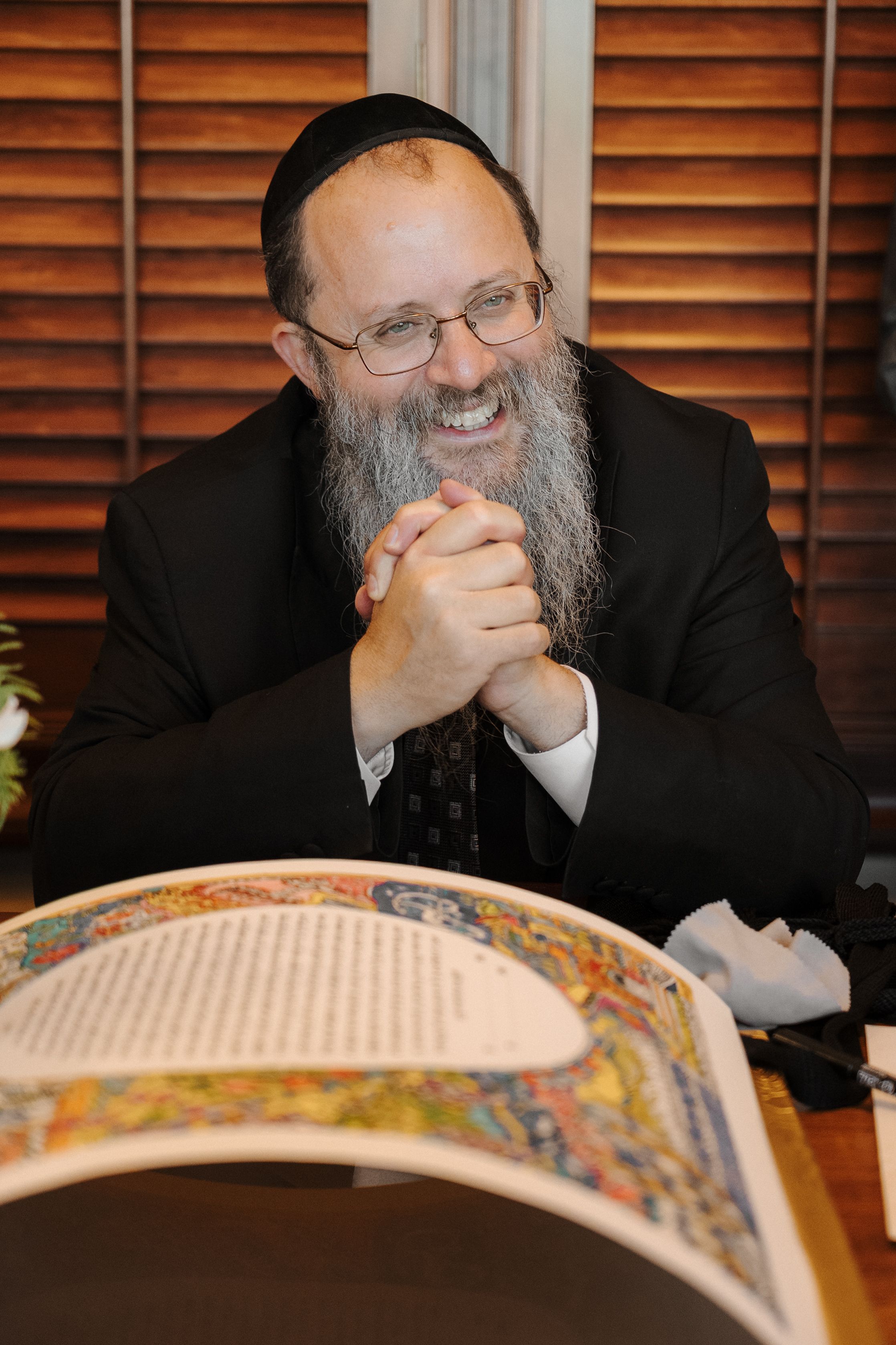Orthodox Jewish man with long gray beard smiles joyfully while studying a traditional religious text at a table.