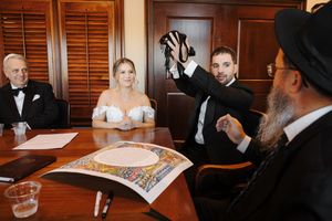 Wedding ceremony participants signing documents at a wooden table in an elegant room with wooden shutters.