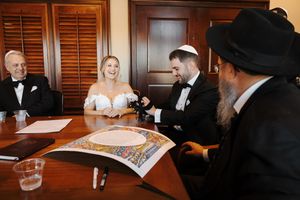 A couple signs their ketubah during a traditional Jewish wedding ceremony while witnesses look on at a wooden table.