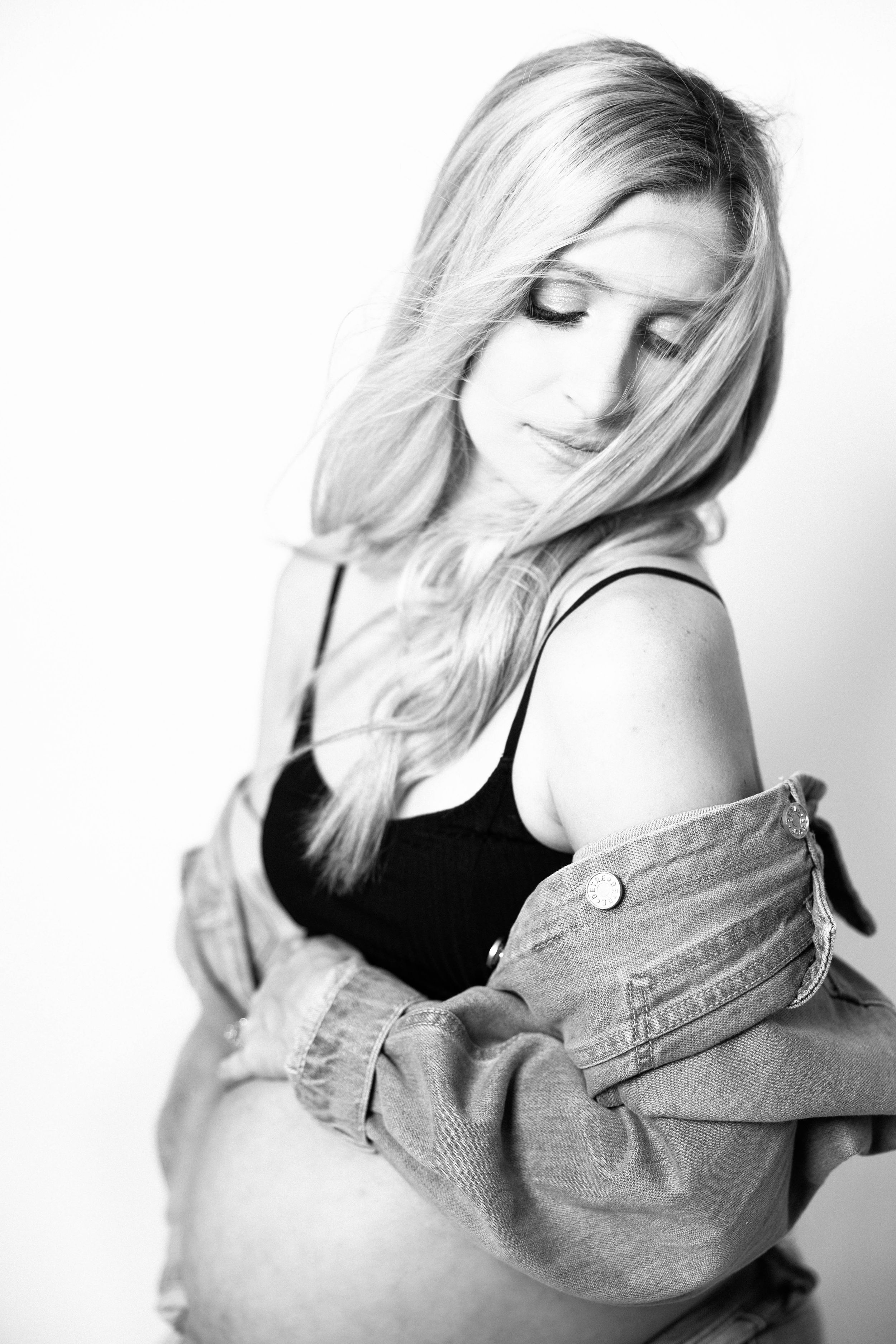 Black and white maternity photo of a blonde woman in denim jacket cradling her pregnant belly softly.