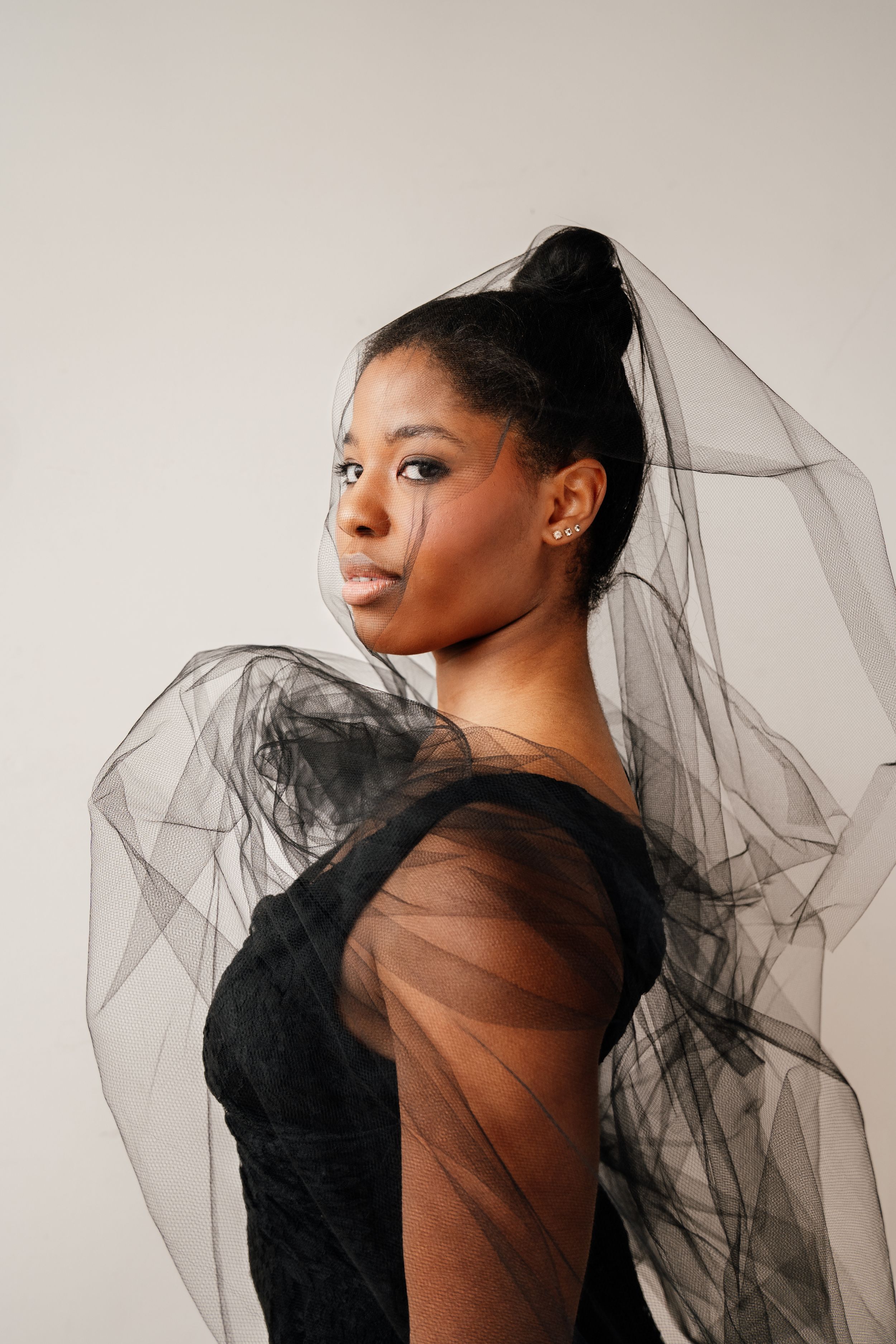 Elegant fashion portrait featuring black tulle dress with sheer sleeves against neutral background.