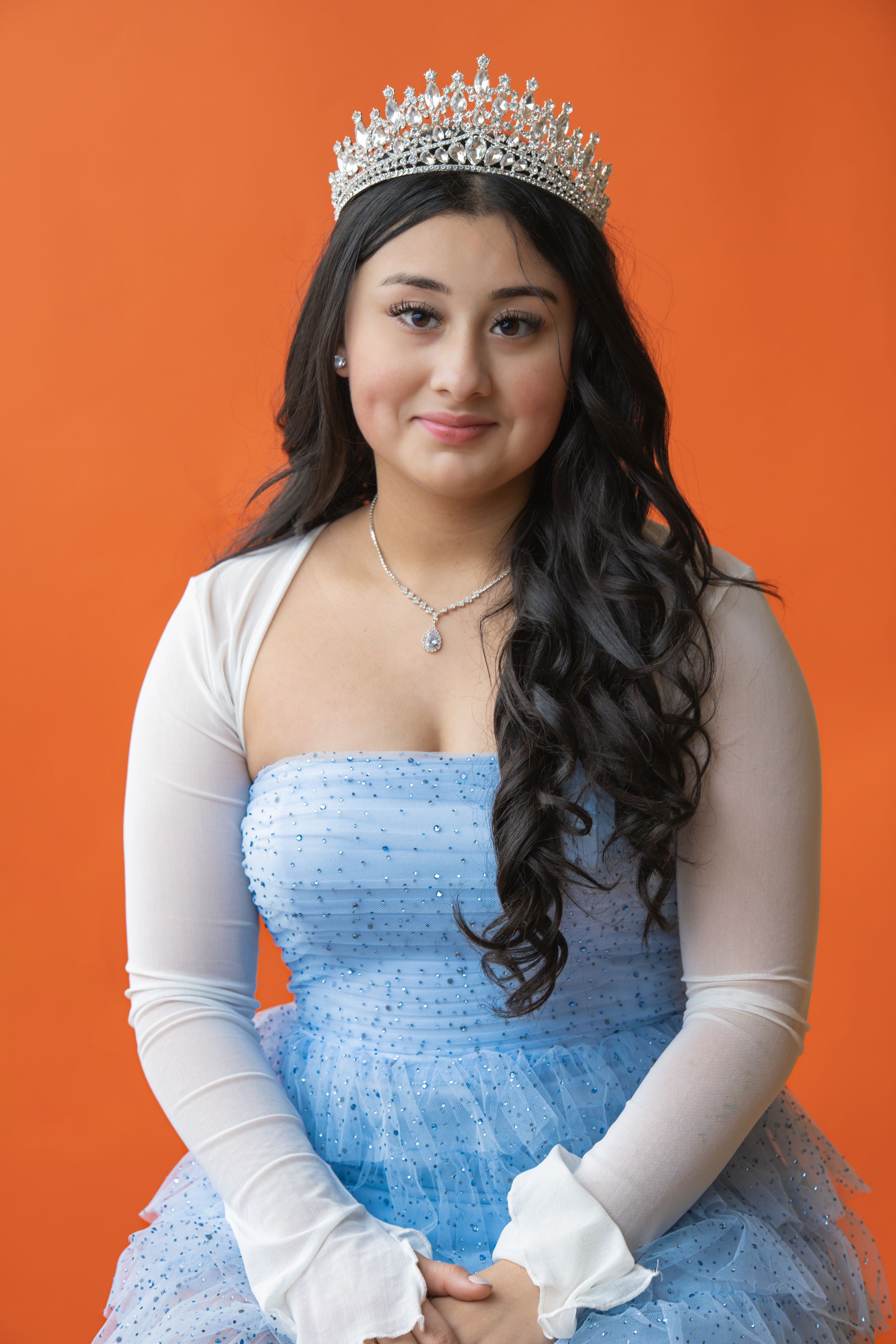 Young woman in blue tulle gown and crystal tiara poses gracefully against vibrant orange background.