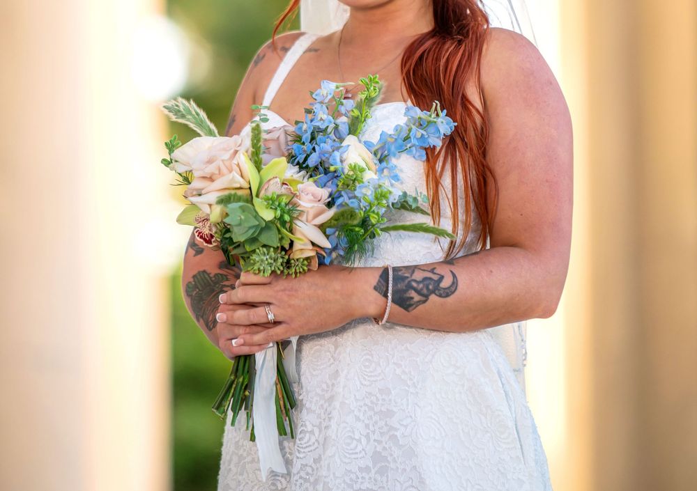 A delicate bridal bouquet with pink and blue flowers is held by tattooed hands against a white lace dress.