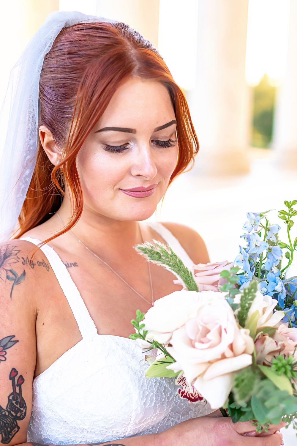 Bride in white lace dress holding delicate bouquet of pink roses and blue flowers with greenery.