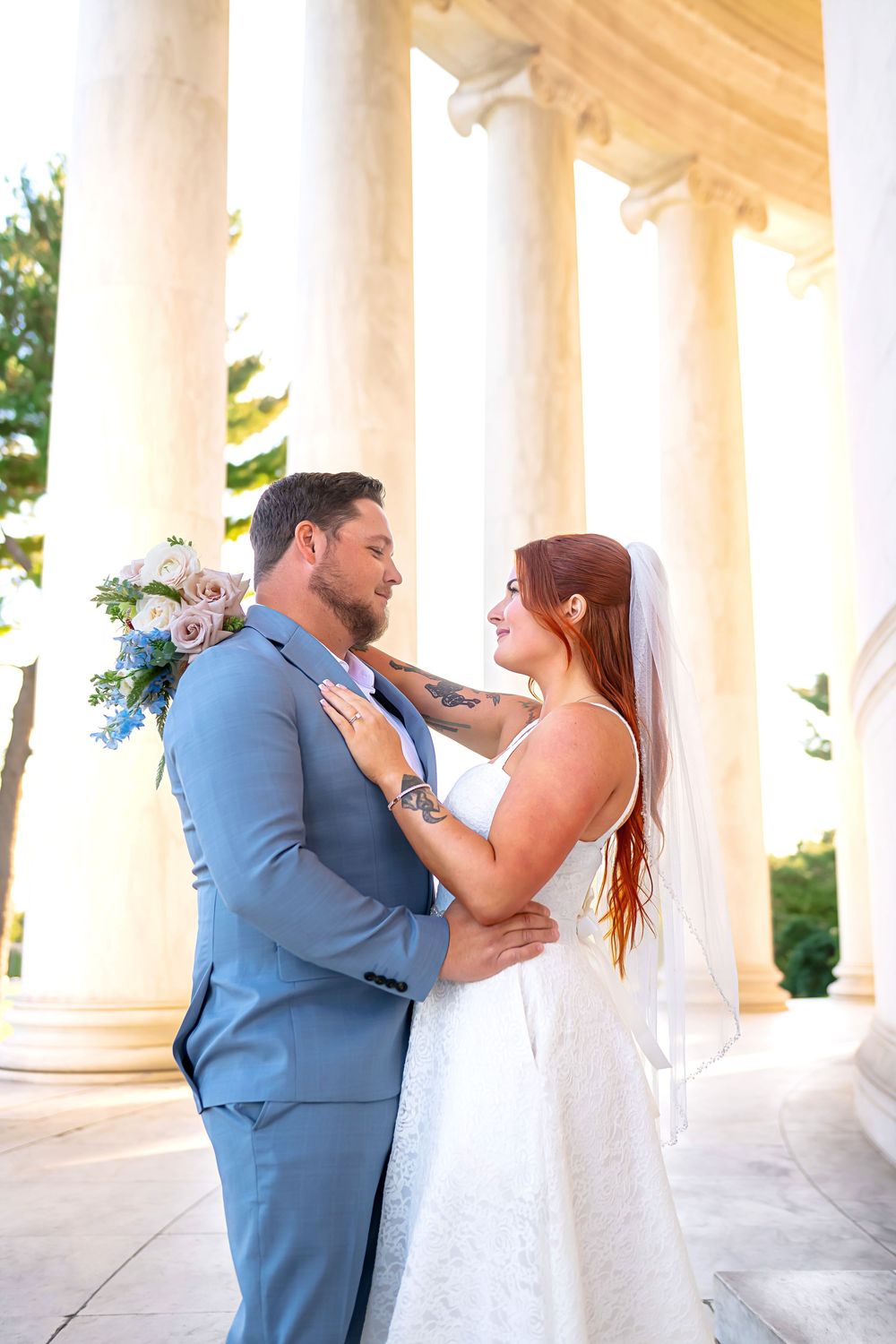 A romantic wedding couple embraces among elegant white columns in a classic architectural setting at sunset.