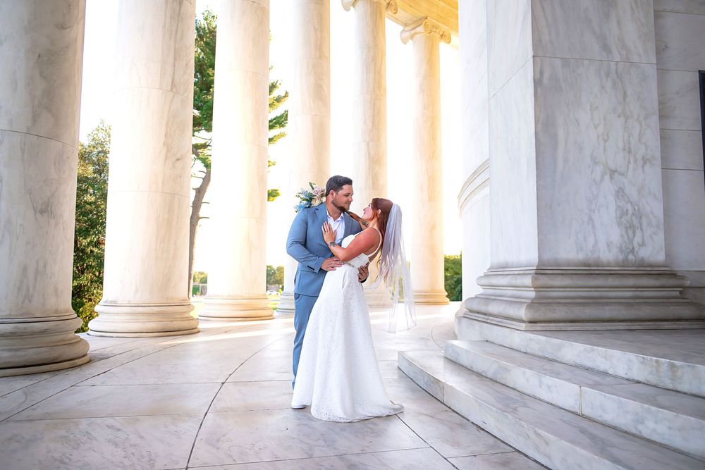A couple in formal wedding attire share a romantic moment between elegant white marble columns at sunset.