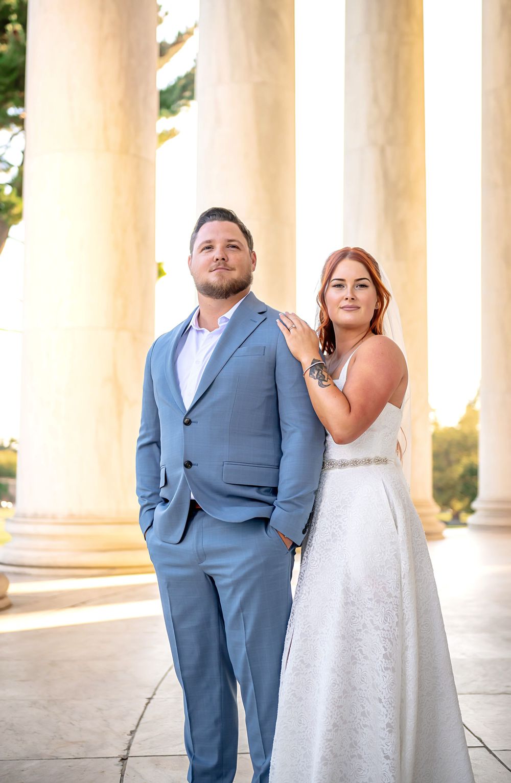 A couple in formal attire poses elegantly between white columns at their wedding photoshoot.