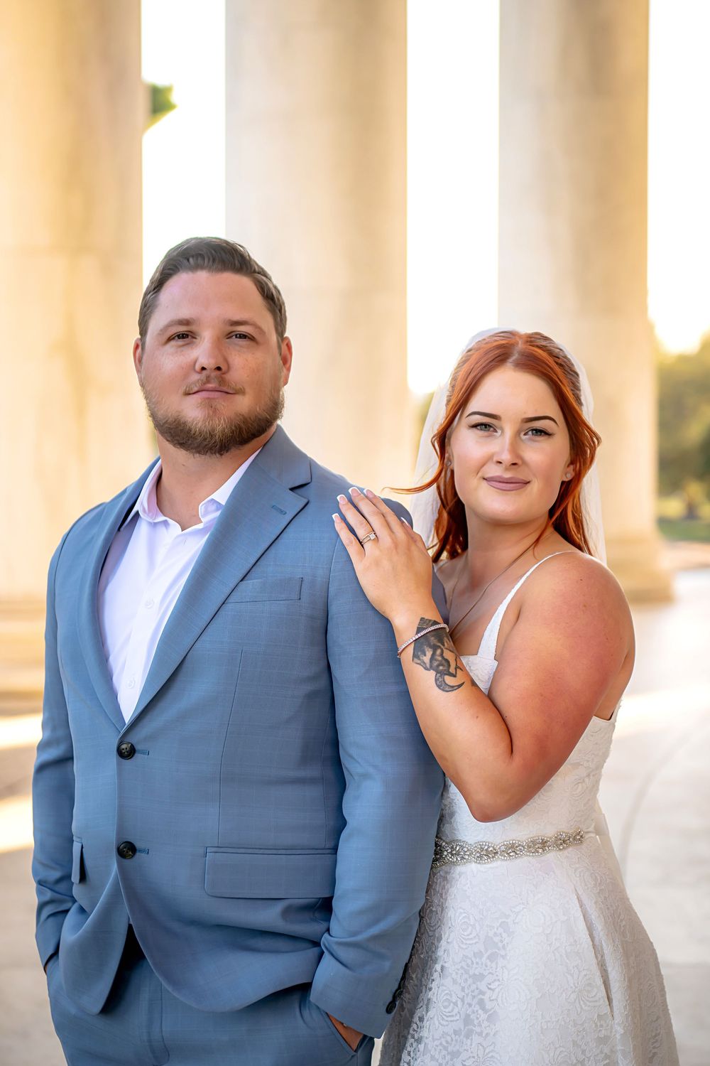 Wedding couple poses against white pillars in formal attire with light blue suits and white dress at sunset.