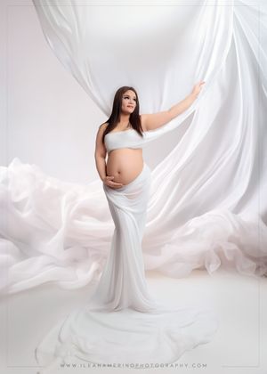Pregnant woman in white flowing fabric gown, dramatic white studio maternity photo.