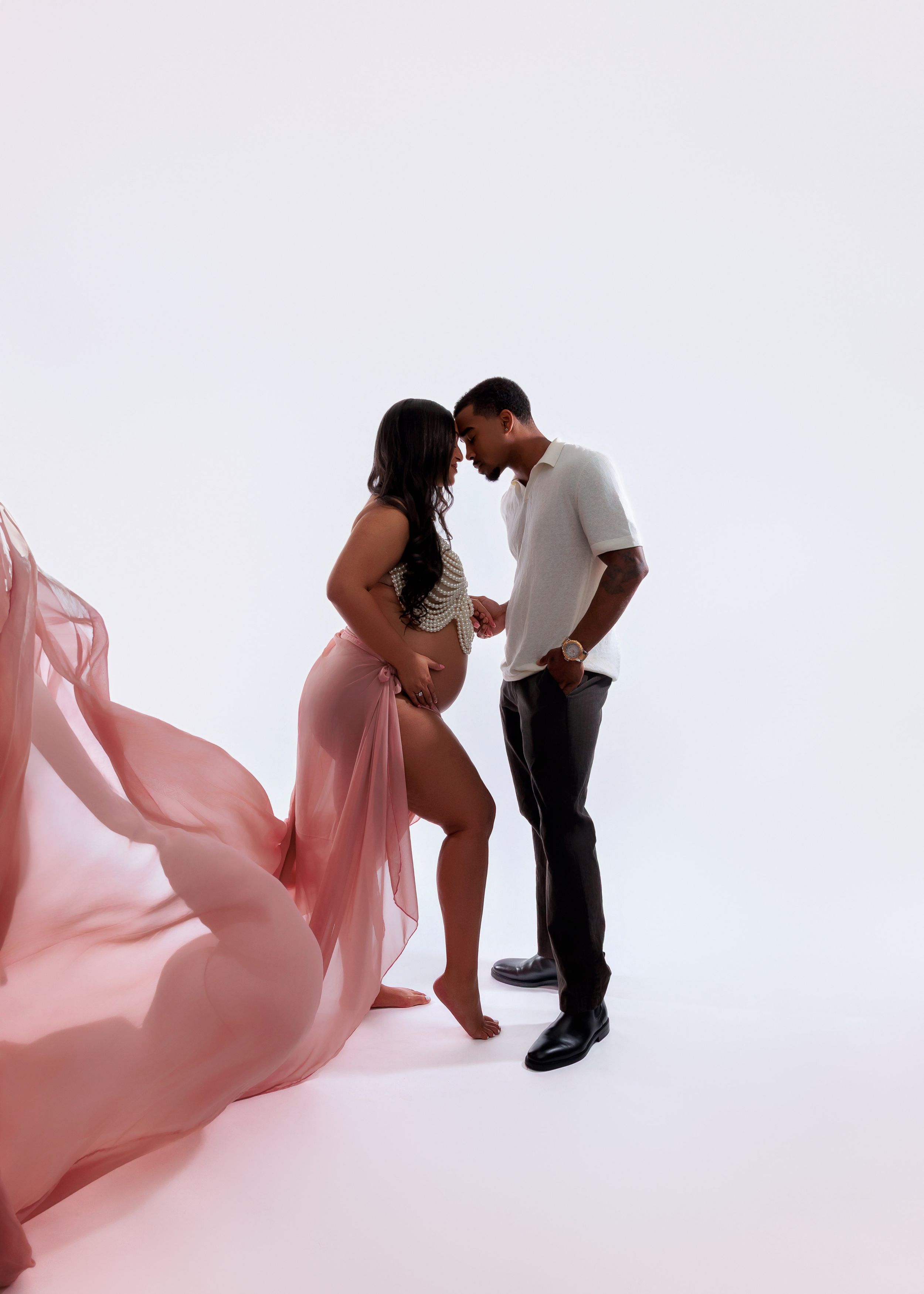 Couple shares tender moment during maternity photoshoot with flowing pink fabric on white backdrop.