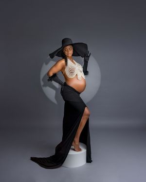 Pregnant woman in black hat, gloves, and skirt poses elegantly on white pedestal.