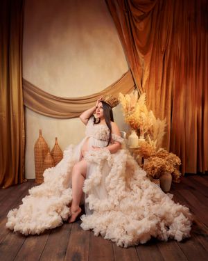 Expectant mother in fluffy cream ruffled gown and crown sits in warm golden studio.
