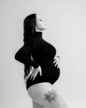 Black and white maternity portrait of pregnant woman in black bodysuit with thigh tattoo.