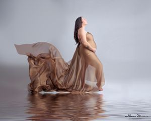 Pregnant woman draped in flowing golden fabric poses near reflective water surface.