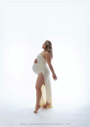 Pregnant woman in white sleeveless gown with thigh slit poses against bright white backdrop.