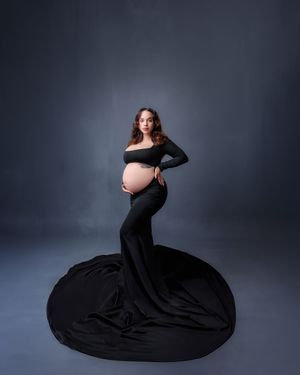 Pregnant woman in black off-shoulder gown with long train poses on dark blue backdrop.
