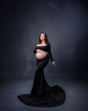 Pregnant woman in elegant black off-shoulder gown with train poses gracefully in studio.