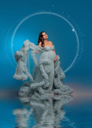 Expectant mother in fluffy blue tulle gown poses before glowing circle on blue backdrop.