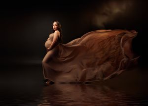 Pregnant woman draped in flowing brown fabric stands in reflective water against dark background.