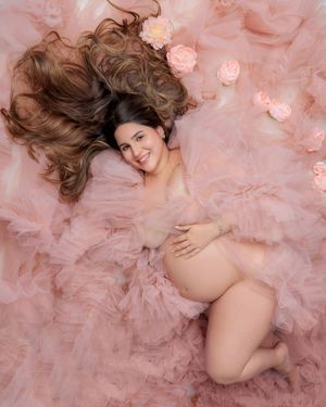 Smiling pregnant woman lies on pink tulle fabric surrounded by roses in overhead maternity shot.