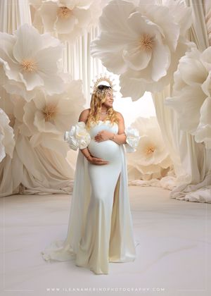 Pregnant woman in ivory gown and floral crown poses before large paper flowers in elegant studio.