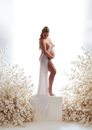 Pregnant woman in sheer white gown cradles bump on pedestal surrounded by white floral arrangements.