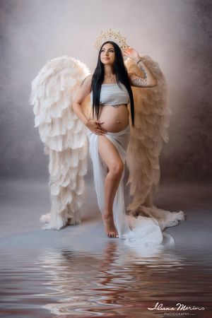 Pregnant woman with large angel wings and crown stands in water in ethereal maternity portrait.