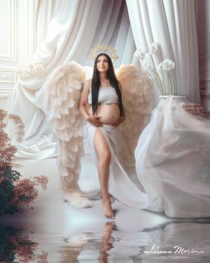 Maternity photo of a woman in white dress with angel wings and gold halo crown in an ethereal studio setting.