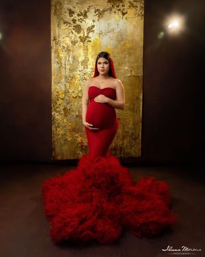 Pregnant woman in elegant red ruffled mermaid gown poses before a golden textured backdrop in a studio.