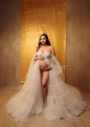 Pregnant woman in sheer ivory tulle gown poses gracefully against a warm golden studio background.