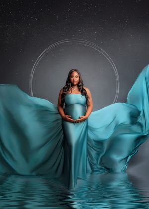 Maternity portrait of a woman in teal flowing fabric gown standing in water under a starry night backdrop.