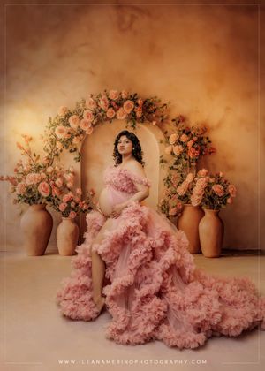 Maternity portrait of a woman in pink ruffled gown seated before a floral arch with roses and terracotta vases.
