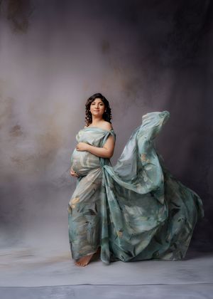 Pregnant woman in sage green floral flowing gown poses elegantly against a soft gray studio background.