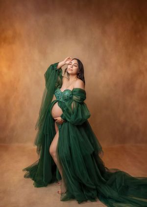 Pregnant woman in elegant emerald green tulle maternity gown posing gracefully in a professional studio photo shoot.
