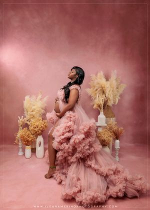 Pregnant woman in pink ruffled tulle gown poses with dried pampas grass arrangements on a pink studio backdrop.