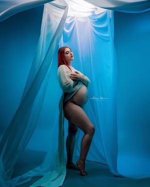 Pregnant woman with red hair poses in sheer teal wrap against vibrant blue backdrop in artistic maternity shoot.