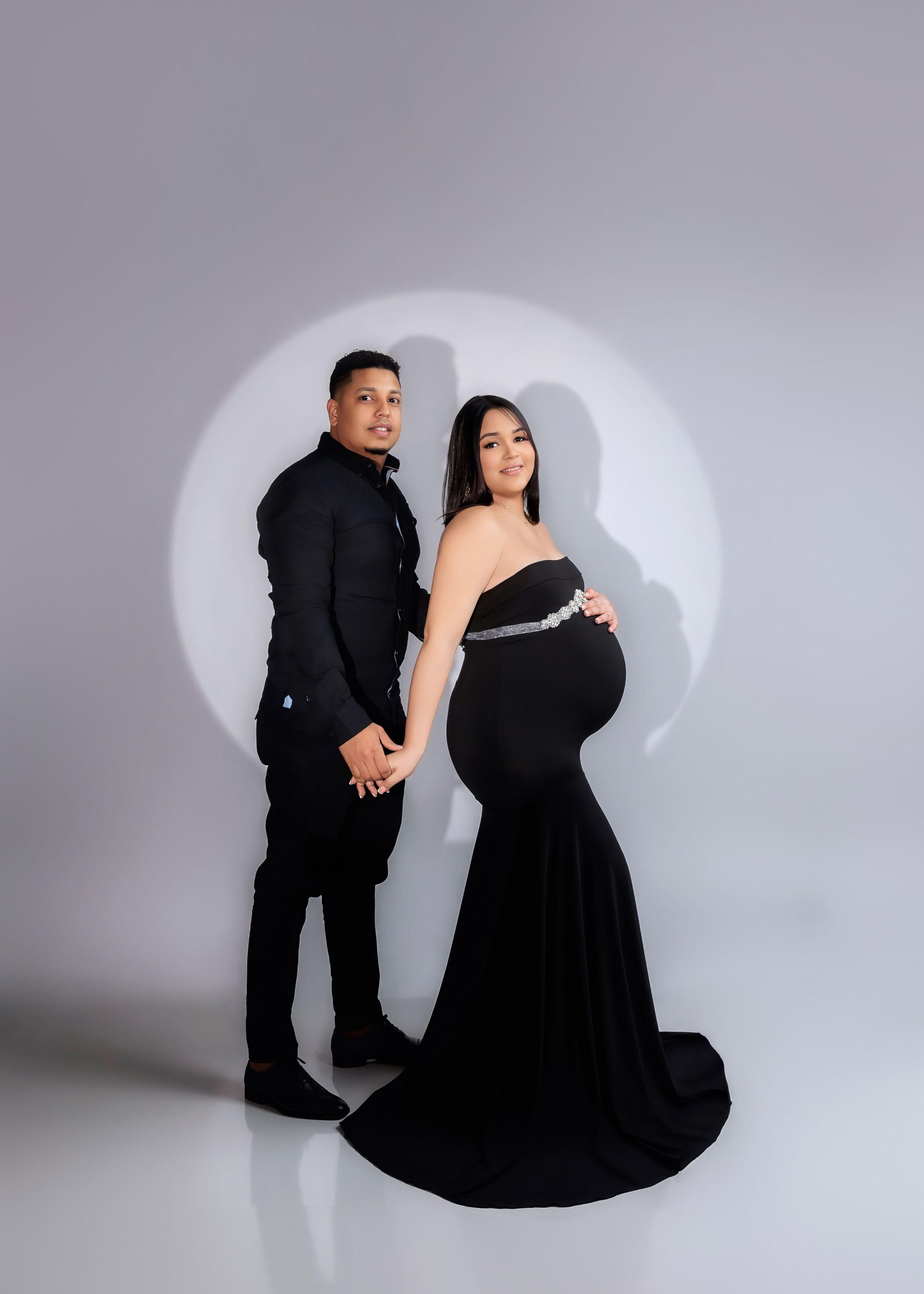 Couple poses elegantly in matching black outfits during maternity photoshoot on gray backdrop.