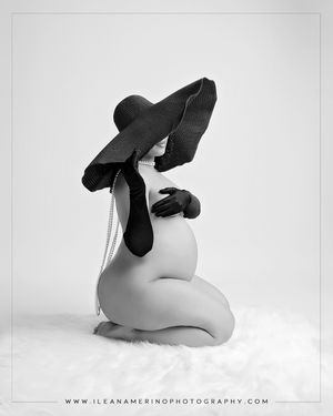 Black and white maternity photo of pregnant woman sitting in wide-brim hat holding baby bump on white fur.