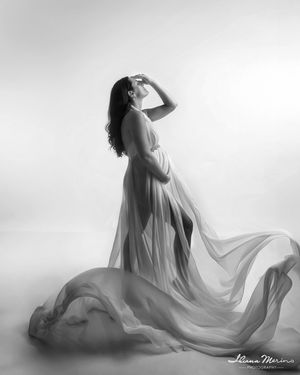 Elegant black and white maternity photo of pregnant woman in flowing sheer gown with dramatic fabric movement.