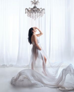 Pregnant woman in sheer white gown poses beneath crystal chandelier in a dreamy, ethereal maternity studio photo.