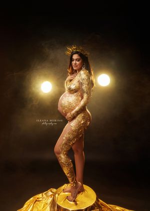 Pregnant woman in gold sequin bodysuit and crown poses dramatically under spotlights in a glamorous maternity shoot.